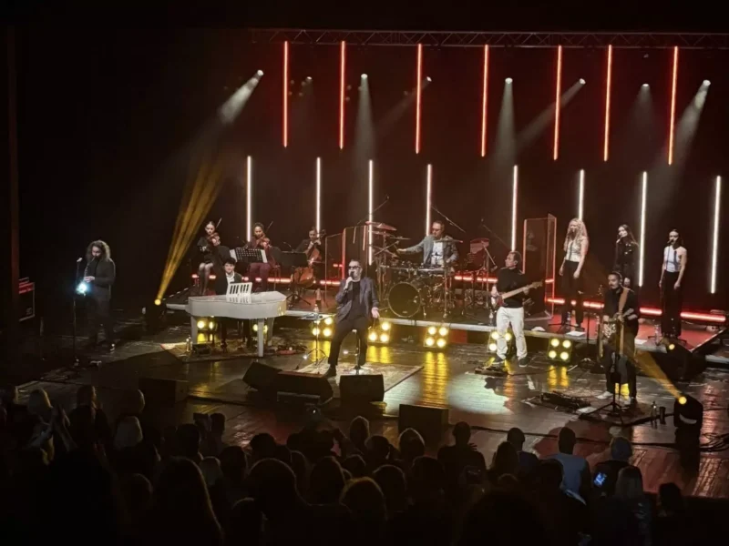 Živá koncertná scéna zahŕňa hudobníkov vystupujúcich na pódiu s farebnými vertikálnymi svetelnými rampami. Publikum je zaujaté a vytvára energickú atmosféru.