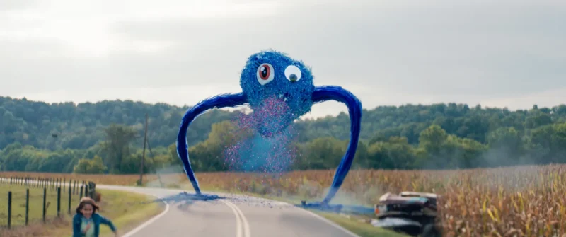 Na vidieckej ceste stojí veľký, chlpatý modrý tvor s kreslenými očami a dlhými končatinami a uvoľňuje farebné častice. Človek uteká, čím vyjadruje naliehavosť.