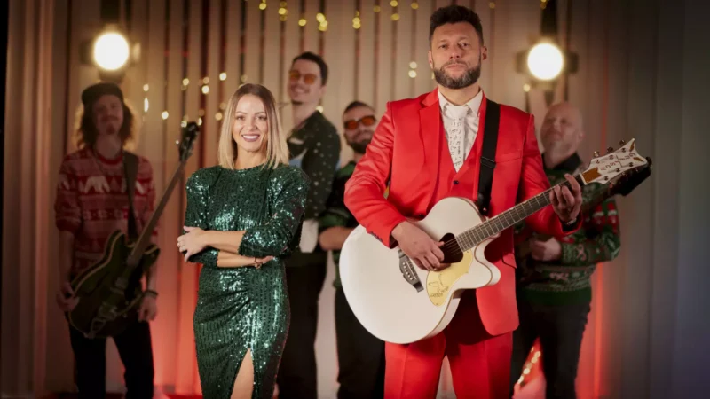 Usmievavá žena v zelených flitrovaných šatách stojí vedľa muža v červenom obleku, ktorý drží gitaru. V pozadí sú štyria členovia kapely vo slávnostnom oblečení a teplé osvetlenie vytvára živú, slávnostnú atmosféru.