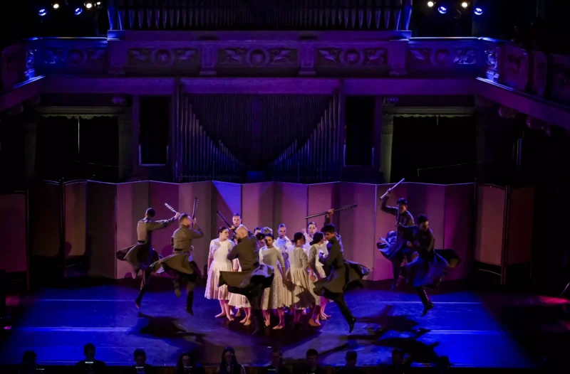 Účinkujúci v tradičných odevoch energicky tancujú na pódiu a držia v rukách paličky. Tlmené osvetlenie a zdobené pozadie vytvárajú dramatickú a dynamickú scénu.