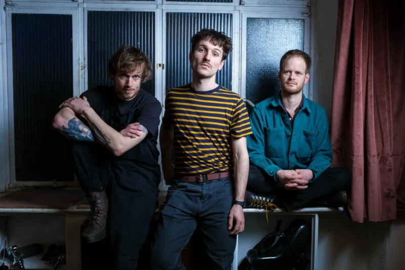 Traja muži ležérne pózujú v interiéri. Jeden sedí s potetovanou rukou opretou o koleno, druhý v pruhovanej košeli sa opiera o pult a tretí sedí so skríženými nohami. Nálada je uvoľnená, s tlmenými tónmi.