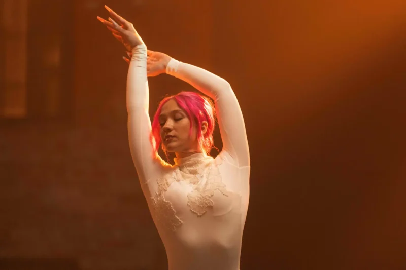 Tanečnica so žiarivo ružovými vlasmi pózuje elegantne so zatvorenými očami v detailne prepracovanom bielom oblečení. Teplé osvetlenie vytvára éterickú a pokojnú atmosféru.