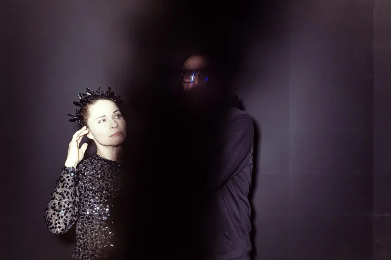 Žena v flitrovaných šatách s ježatými vlasmi vyzerá zamyslene a dotýka sa ucha, zatiaľ čo tieňovaná postava s okuliarmi stojí čiastočne zakrytá. Tmavý, tajomný tón.