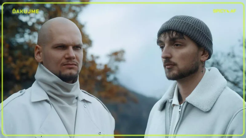 Dvaja muži v zimnom oblečení stoja vonku uprostred jesenného lístia. Jeden je plešatý s vážnym výrazom, druhý v čiapke vyzerá zamyslene.