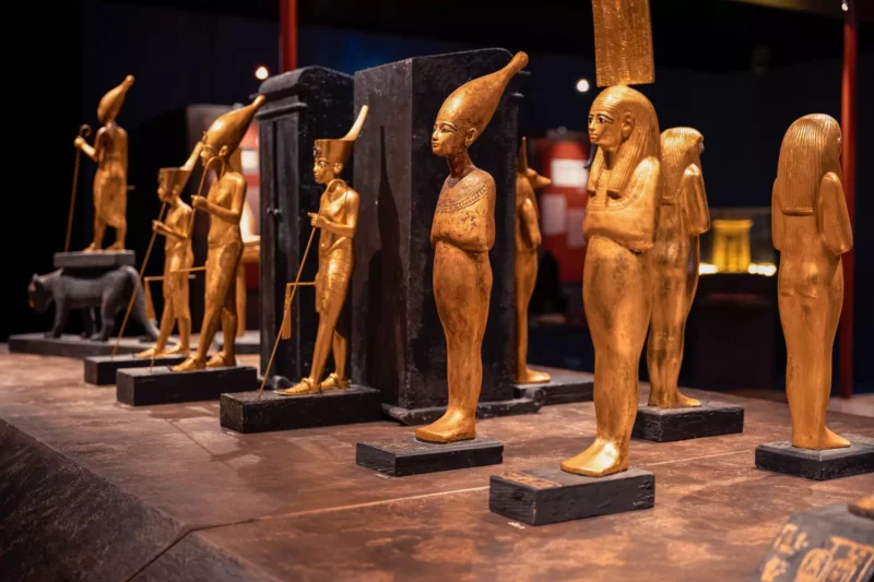 Zlaté sochy staroegyptských postáv, každá stojaca na čiernych podstavcoch, sú vystavené v slabo osvetlenom múzejnom prostredí a evokujú pocit tajomstva a starobylosti.