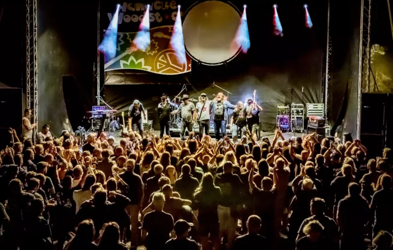 Kapela energicky vystupuje na slabo osvetlenom pódiu, osvetlenom žiarivými reflektormi. K pódiu čelí veľký, nadšený dav s rukami zdvihnutými v potlesku.