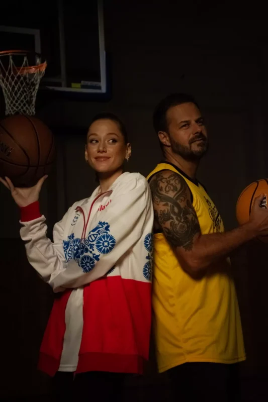 Žena v červeno-bielom saku a muž v žltom tielku stoja chrbtom k sebe, obaja držia basketbalovú loptu. V pozadí je viditeľný basketbalový kôš.