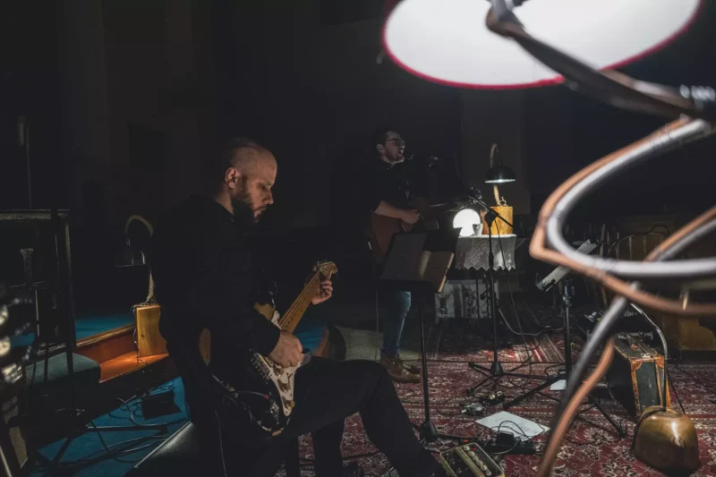 Malá kapela vystupuje v slabo osvetlenej miestnosti s útulnou, intímnou atmosférou. Gitarista hrá na elektrickú gitaru, zatiaľ čo iný hudobník spieva do mikrofónu.