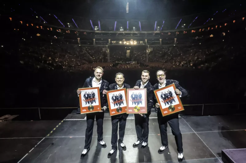 Na pódiu stoja štyria muži, usmievajú sa a držia zarámované zlaté platne. Veľký dav v slabo osvetlenej aréne v pozadí tlieska a vytvára oslavnú atmosféru.