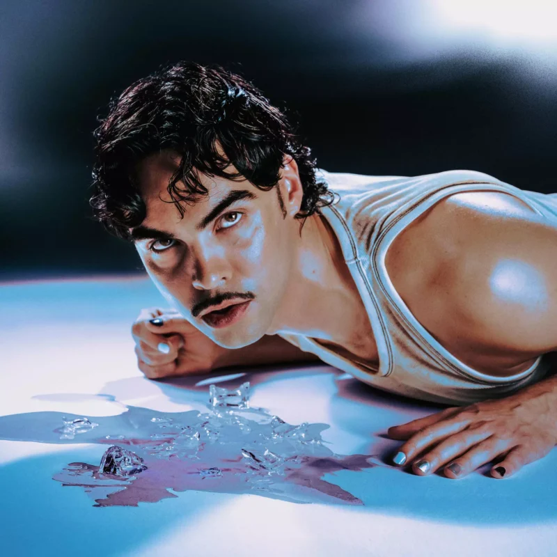 Na podlahe pokrytej topiacim sa ľadom leží osoba s tmavými vlasmi a fúzikmi. Má na sebe tričko bez rukávov, nalakované nechty a uprene sa pozerá.