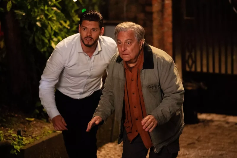 Dvaja muži, jeden v bielej košeli a druhý v bunde, sa znepokojene krčia v slabo osvetlenej uličke. Scéna vyjadruje napätie a naliehavosť.