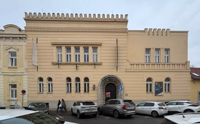 Historická béžová budova s ​​klenutými oknami a cimbuřím v tvare strechy. Pred ňou parkuje niekoľko áut a po chodníku kráčajú dvaja ľudia.