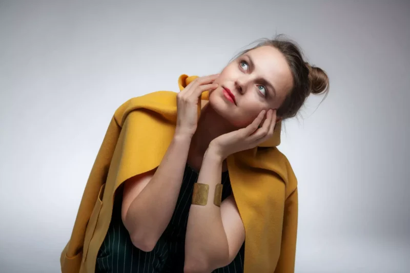 Žena v žltom kabáte vyzerá zamyslene, ruky má položené na tvári. Jej výraz je zamyslený na neutrálnom pozadí, čo vyžaruje pocit introspekcie.