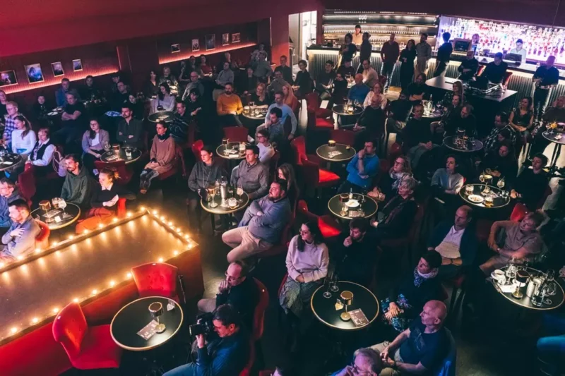 Veľký dav sedí za okrúhlymi stolmi v slabo osvetlenom priestore s červenými stoličkami. Ľudia čelia pódiu osvetlenému teplými reťazovými svetlami, čo naznačuje uvoľnenú a pozornú atmosféru.