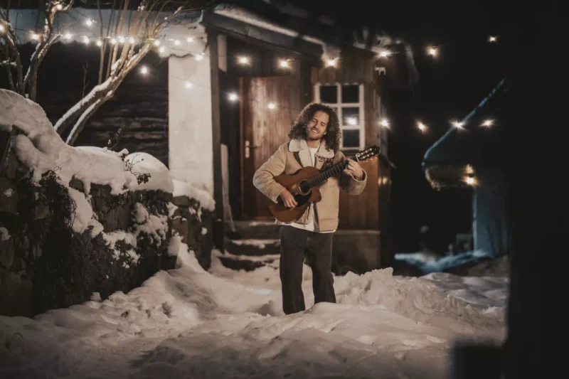 Dlhovlasý muž hrá v noci vonku na gitare. Stoja na zasneženej zemi blízko príjemne osvetlenej chatky, obklopená svetelnými reťazami. Scéna je útulná a pokojná.