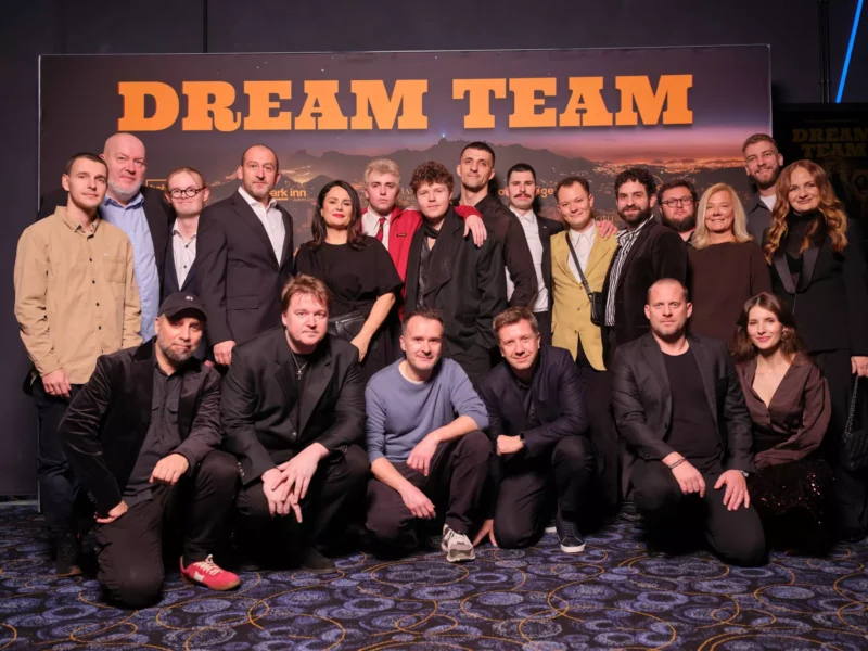 Veľká skupina ľudí pózuje pred kulisou „Dream Teamu“. Vyzerajú veselo a sú oblečení v poloformálnom až ležérnom oblečení.