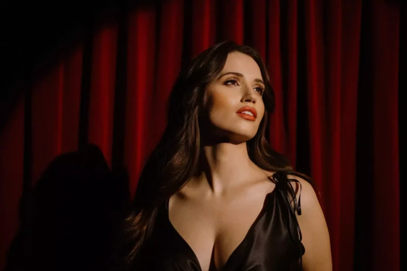 Žena v čiernych šatách je osvetlená na pozadí tmavočervenej opony. Hľadí hore a v náladovom osvetlení vyžaruje sebavedomie a eleganciu.