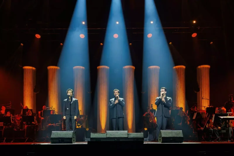 Traja umelci v oblekoch spievajú na pódiu pod modrými reflektormi, za nimi sú žiariace oranžové stĺpy. V tlmenom pozadí je viditeľný orchester, ktorý vytvára elegantnú atmosféru.