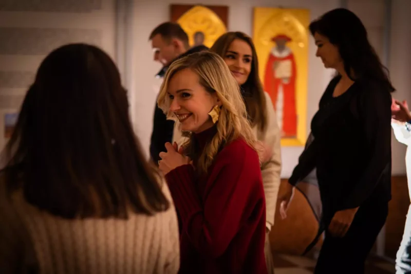 Skupina ľudí sa socializuje v interiéri, jedna žena v červenom svetri sa srdečne usmieva. Pozadie zdobia farebné umelecké diela, ktoré vytvárajú živú a veselú atmosféru.