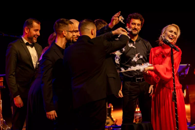 Žena v červených šatách spieva do mikrofónu, zatiaľ čo skupina formálne oblečených mužov okolo nej na pódiu radostne vypúšťa pierka.