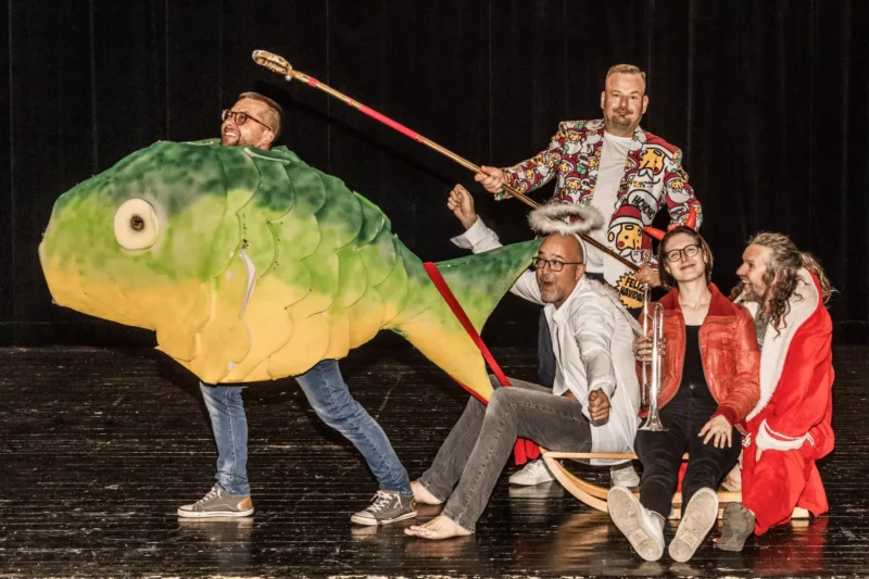 Skupina ľudí na javisku; jeden vo veľkom kostýme zelenej ryby, ostatní sedia na saniach vo farebných outfitoch. Nálada je hravá a rozmarná.
