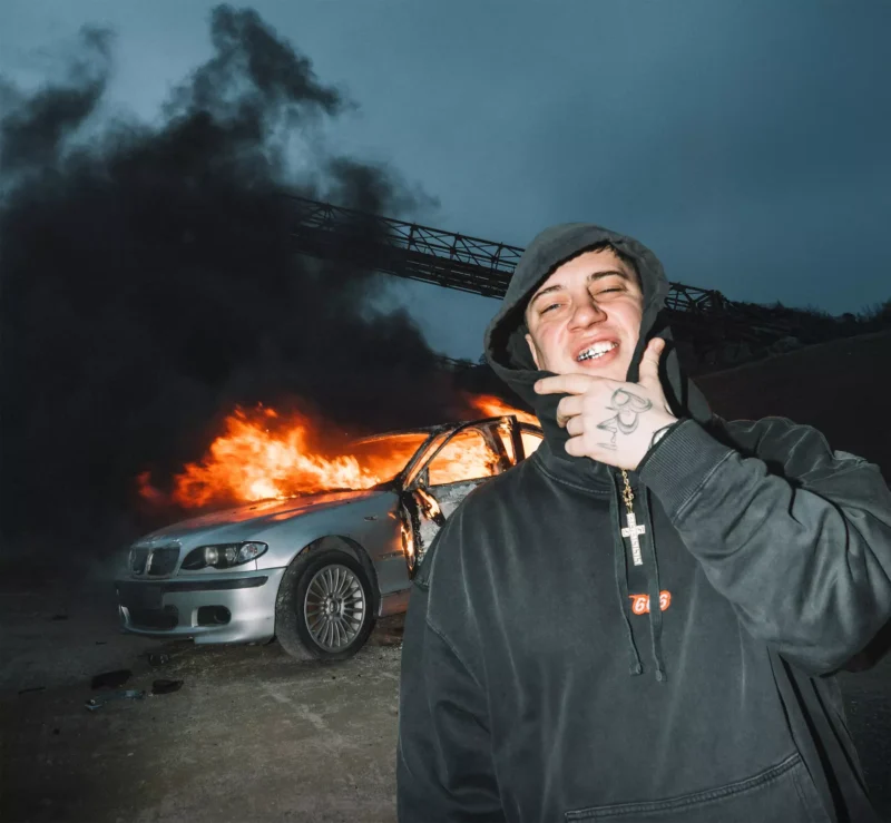 Osoba v mikine s kapucňou sa usmieva s rukou na brade, zatiaľ čo v pozadí horí strieborné auto, ktoré pod pochmúrnou oblohou vypúšťa hustý čierny dym.