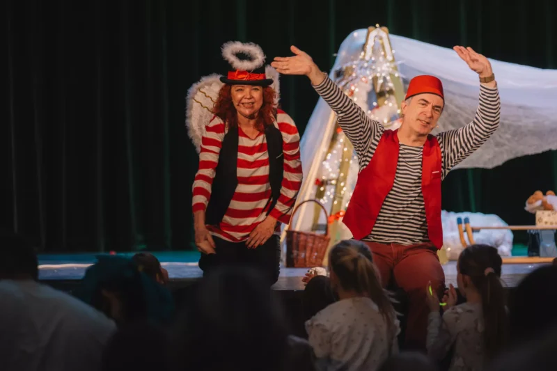 Dvaja účinkujúci vo slávnostných kostýmoch, jeden s krídlami a svätožiarou, sa usmievajú a gestikulujú smerom k detskému publiku na javisku. Pozadie tvorí útulná, vyzdobená scéna.