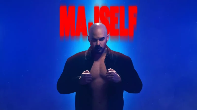 Plešatý muž stojí sebavedomo na modrom pozadí v rozopnutom tmavom saku. Nad jeho hlavou je zobrazený tučný červený nápis „MAJSELF“.