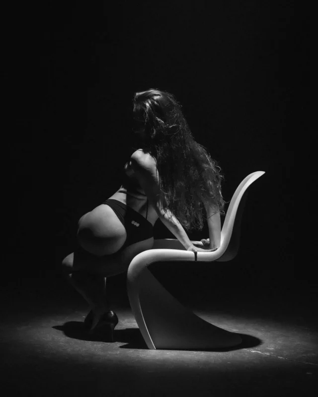 Monochromatický obraz ženy s dlhými vlasmi pózujúcej na modernej, zakrivenej stoličke v slabo osvetlenom prostredí. Vyžaruje tajomnú a dramatickú atmosféru.