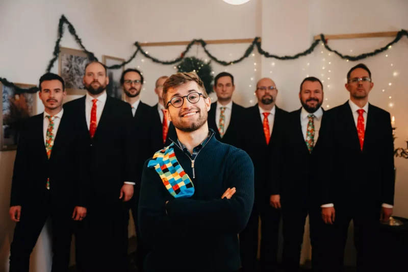 Usmievavý muž v modrom svetri s farebnou šatkou sebavedomo stojí pred skupinou formálne oblečených mužov v oblekoch so slávnostnými kravatami. Miestnosť je vyzdobená girlandami a trblietavými svetielkami, ktoré vytvárajú veselú sviatočnú atmosféru.