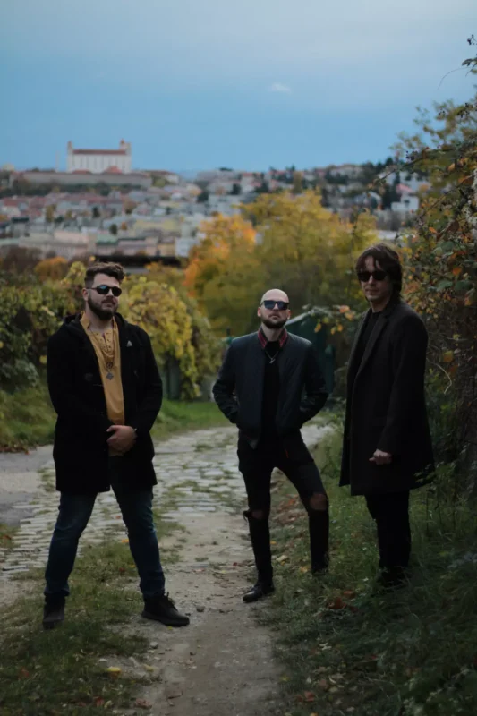 Traja muži v tmavých kabátoch a slnečných okuliaroch stoja na rustikálnom chodníku obklopenom jesenným lístím. V diaľke sa objavuje mestská krajina a hrad.