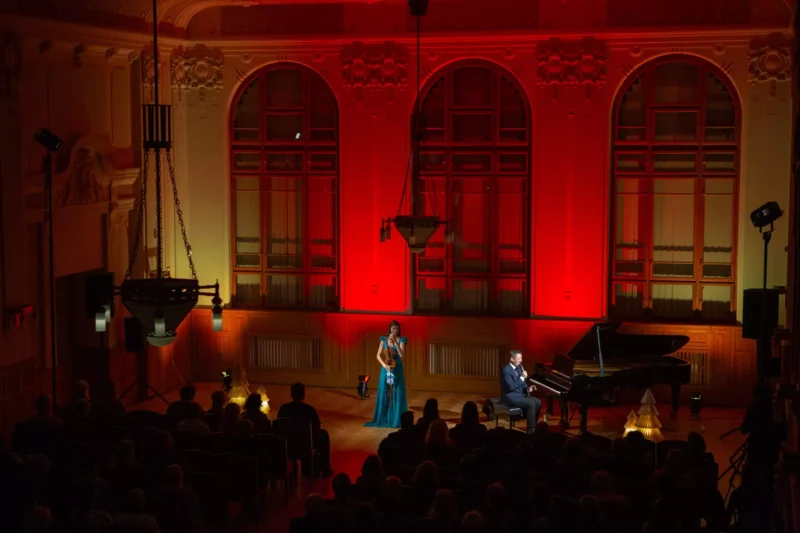 Huslistka v modrých šatách hrá vedľa sediaceho klaviristu na klavíri v elegantnej sále. Červené osvetlenie vytvára teplú a dramatickú atmosféru.