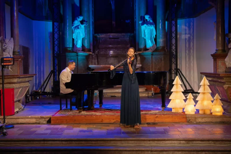 Žena v splývavých tmavomodrých šatách vášnivo hrá na husle vedľa klaviristu na klavíri. Javisko zdobia papierové lampy v tvare stromčeka, ktoré sú umiestnené na pozadí osvetlených sôch a vytvárajú pokojnú a elegantnú atmosféru.