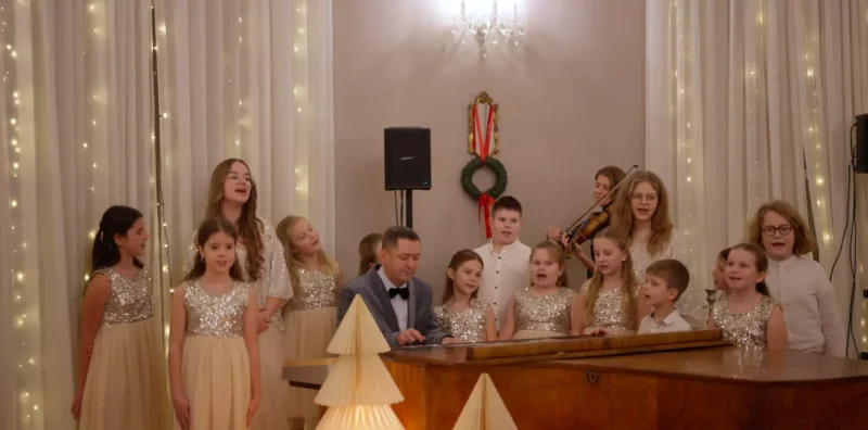 Radostný detský zbor v trblietavých šatách spieva okolo klaviristu na slávnostnom podujatí. Teplé svetielka a veniec vytvárajú veselú sviatočnú atmosféru.