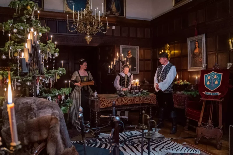 Slabo osvetlená, starožitná miestnosť ozdobená sviečkami a bohato vyzdobeným vianočným stromčekom. Traja ľudia v dobových kostýmoch sa rozprávajú a vytvárajú elegantnú historickú atmosféru.