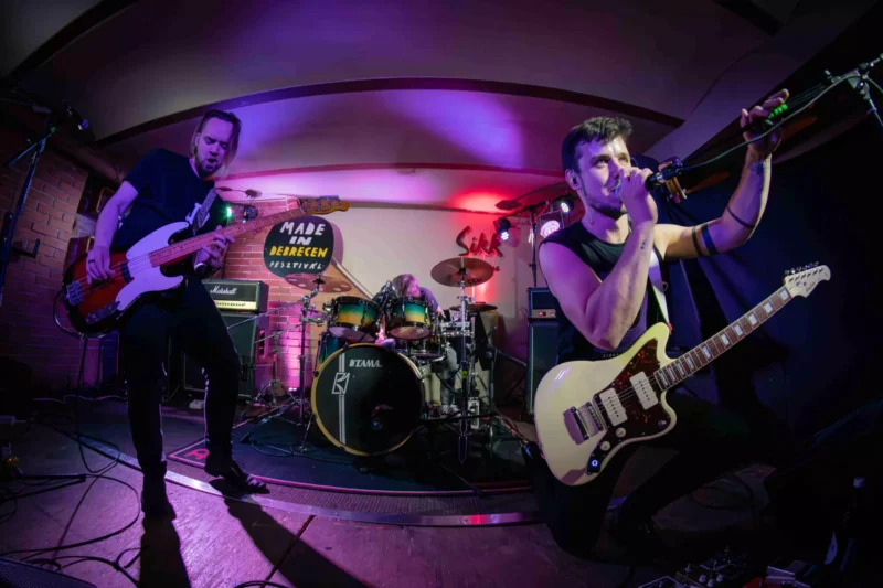 Rocková kapela energicky vystupuje na pódiu s žiarivým fialovým a červeným osvetlením. Gitarista spieva do mikrofónu, zatiaľ čo basgitarista a bubeník hrajú vášnivo.