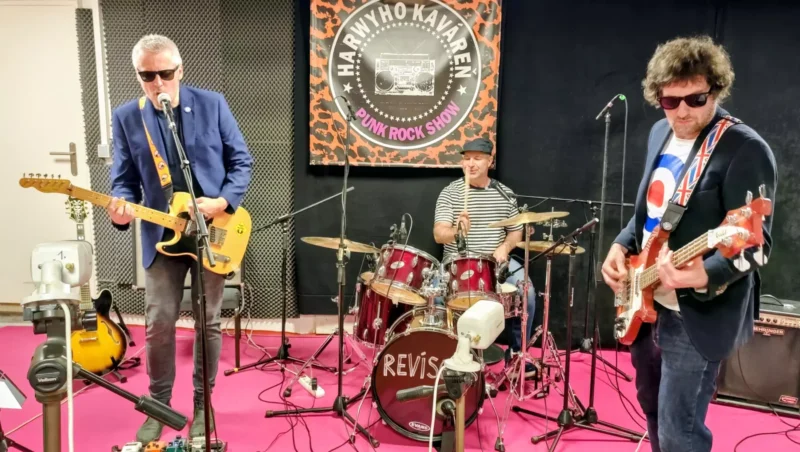 Traja hudobníci energicky vystupujú na ružovom pódiu. Vpredu hrajú gitarista a basgitarista, v pozadí bubeník. Za nimi visí punk rockový transparent.