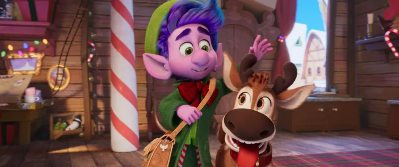 Animovaný elf s fialovými vlasmi a zeleným oblečením sa usmieva vedľa šťastného soba vo vnútri slávnostnej dielne a vytvára tak veselú sviatočnú atmosféru.