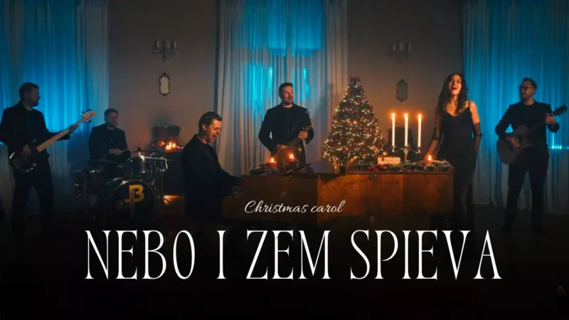 Kapela hrá vianočnú koledu v slabo osvetlenej miestnosti. Hudobníci hrajú na hudobné nástroje blízko ozdobeného stromčeka a sviečok, čím vytvárajú slávnostnú a príjemnú atmosféru.