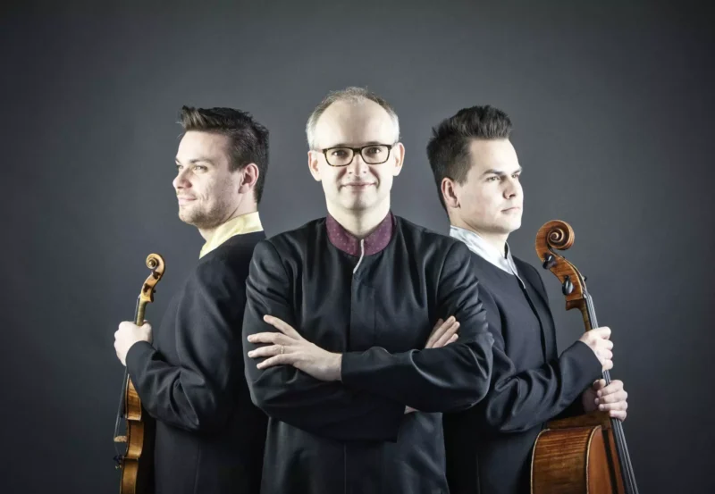 Trio hudobníkov sebavedomo pózuje na tmavom pozadí. Centrálna postava má prekrížené ruky, po stranách sú dvaja muži držiaci husle a violončelo.