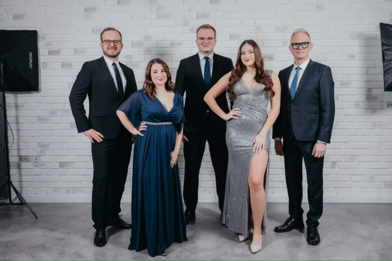 Päť ľudí sebavedomo pózuje vo formálnom oblečení pri bielej tehlovej stene. Dve ženy majú na sebe elegantné šaty a traja muži sú v oblekoch, z čoho vyžaruje slávnostná nálada.