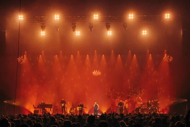 Hudobníci vystupujú na pódiu pod teplým oranžovým osvetlením s lustrami nad hlavou. Veľký dav sleduje vystúpenia a vytvára energickú a živú atmosféru.