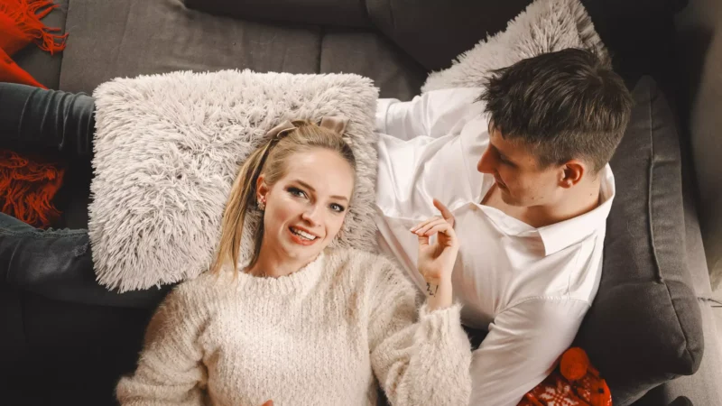 Usmievavá žena v útulnom svetri a muž v bielej košeli relaxujú na sivom gauči s nadýchanými vankúšmi. Atmosféra je vrúcna a intímna.