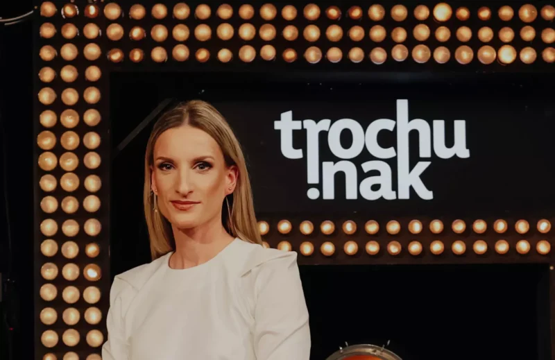 Žena v bielom oblečení stojí sebavedomo v štúdiu s pozadiom zarámovaných svetiel a nápisom „trochu inak“ za ňou. Atmosféra je profesionálna.