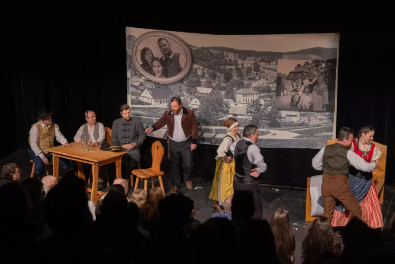 Divadelná scéna s hercami v historických kostýmoch na javisku; v pozadí je stará dedinská fotografia. Diváci pozorne sledujú.