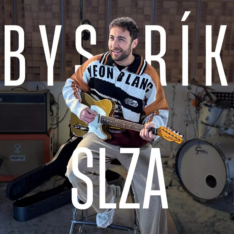 Muž sa usmieva a hrá na zlatej elektrickej gitare v hudobnom štúdiu. Má na sebe farebný sveter; v pozadí sú viditeľné bicie a zosilňovače. Text: „Bystrík SLZA.“