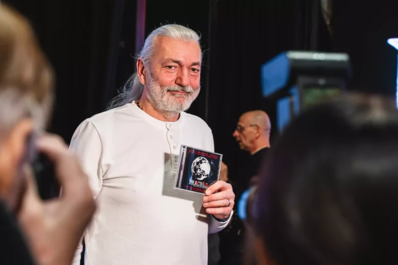 Starší muž v bielej košeli drží CD s filmom „Dracula“ a vrúcne sa usmieva. Fotografi ho obklopujú a zachytávajú tento okamih v slabo osvetlenej miestnosti.