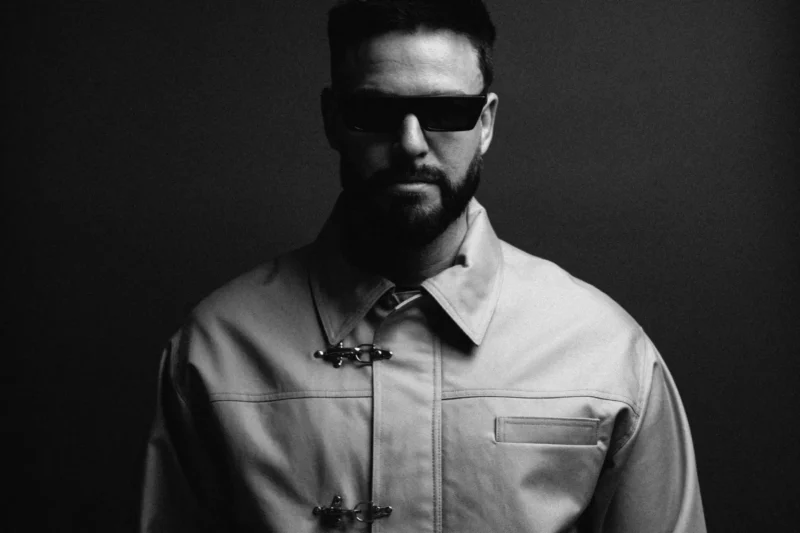 Muž s bradou a slnečnými okuliarmi v zapnutom kabáte sebavedomo stojaci na tmavom pozadí. Tón je tajomný a štýlový.