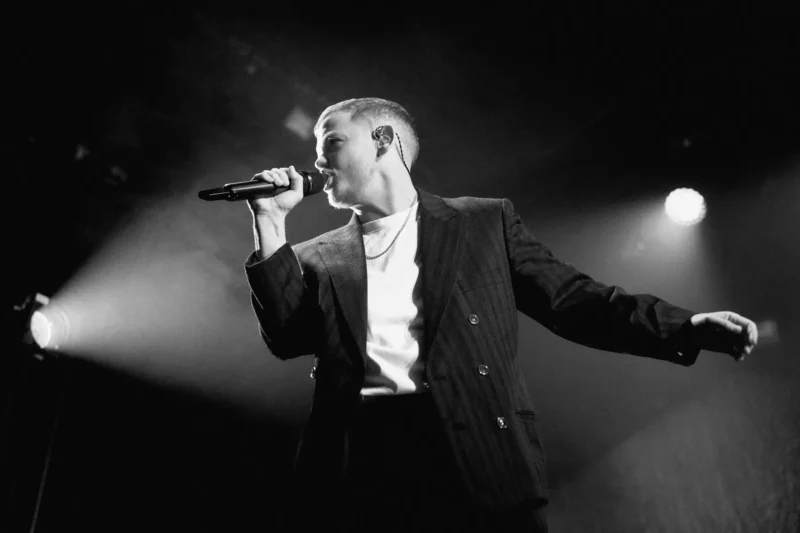 Čiernobiely obraz osoby v obleku, ktorá vášnivo spieva na pódiu. Drží mikrofón pod dramatickým osvetlením, čo vytvára intenzívnu atmosféru vystúpenia.