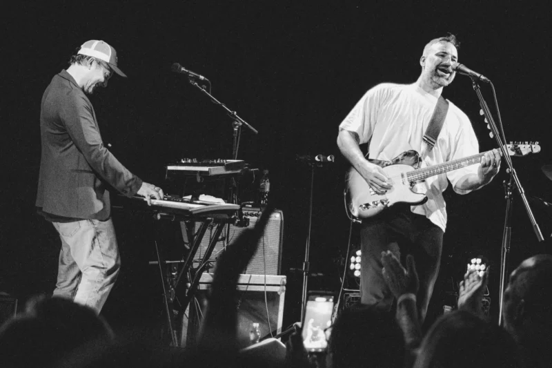 Čiernobiela fotografia kapely hrajúcej na pódiu. Muž v čiapke hrá na klávesy vľavo, zatiaľ čo iný vpravo energicky spieva a hrá na gitare.